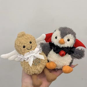 May include: A plush peanut-shaped toy with white wings and a white bow, and a plush penguin toy wearing a red cape and a black bow tie with a bell.