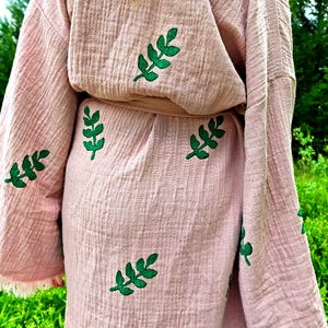 May include: A light pink robe made of textured muslin fabric featuring a repeating pattern of embroidered dark green leaf sprigs. The garment includes long sleeves and a matching fabric tie belt at the waist, showcasing a soft, crinkled finish.