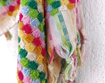 May include: A textile featuring a textured, multi-colored pom-pom pattern in shades of pink, yellow, green, and light blue. The fabric includes a section with a striped weave and decorative knotted fringe tassels along the bottom edge.
