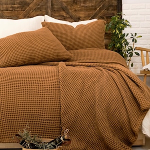 Op de afbeelding: Een tabaksbruine sprei met wafelstructuur en bijpassende kussenslopen op een bed. Het beddengoed heeft een opvallende rasterachtige textuur. Een kleine gevlochten mand met groen en een boek met de titel A Tree in the House staan in de buurt.