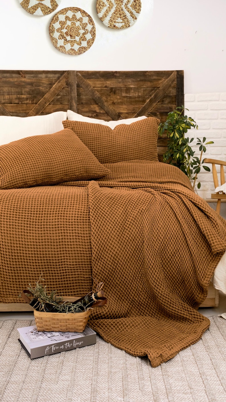 Op de afbeelding: Een tabaksbruine sprei met wafelstructuur en bijpassende kussenslopen op een bed. Het beddengoed heeft een opvallende rasterachtige textuur. Een kleine gevlochten mand met groen en een boek met de titel A Tree in the House staan in de buurt.