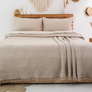 May include: A beige textured bedspread with a fringed edge draped over a bed. The set includes two matching rectangular pillow shams in the same light tan color, featuring a crinkled fabric surface that covers the mattress and pillows.