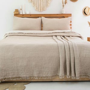 Peut inclure: Un couvre-lit beige texturé avec un bord à franges et deux taies d'oreiller rectangulaires assorties. Le tissu présente un motif de tissage en grille visible, drapé sur un cadre de lit avec une tête de lit en bois, formant une parure.