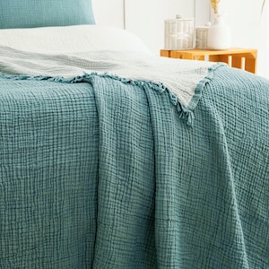 May include: A teal blue textured muslin bedspread with a fringed edge draped over a bed. The bedding set includes a matching teal blue pillowcase and a light gray secondary blanket layered on top, featuring a crinkled, multi-layered fabric weave.