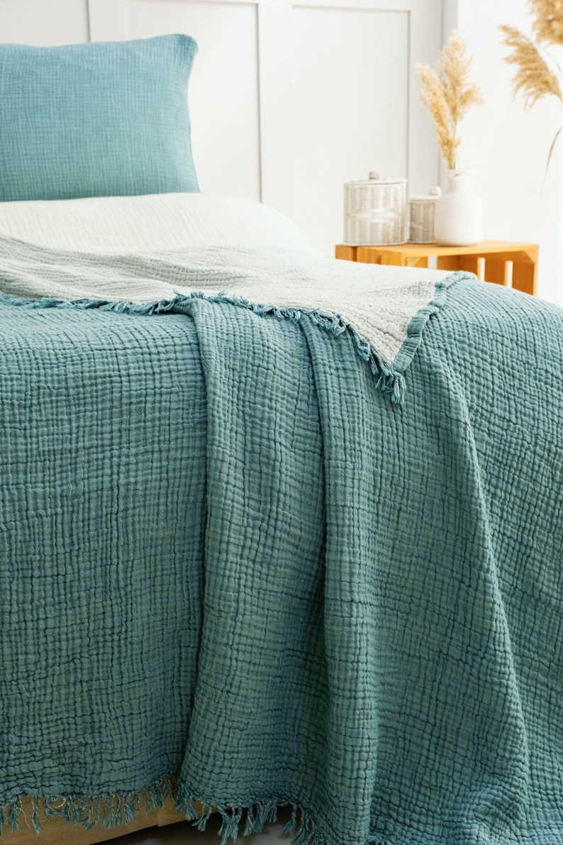 May include: A teal blue textured muslin bedspread with a fringed edge draped over a bed. The bedding set includes a matching teal blue pillowcase and a light gray secondary blanket layered on top, featuring a crinkled, multi-layered fabric weave.