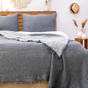May include: A charcoal gray bedding set featuring two rectangular pillows and a large throw blanket with a crinkled, textured fabric finish. The blanket edges are finished with a frayed fringe detail, providing a layered look over the bed.