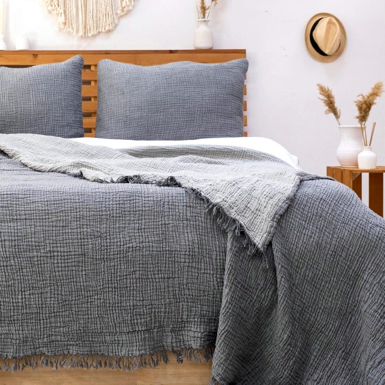 May include: A charcoal gray bedding set featuring two rectangular pillows and a large throw blanket with a crinkled, textured fabric finish. The blanket edges are finished with a frayed fringe detail, providing a layered look over the bed.