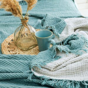May include: A teal and white crinkled muslin throw blanket with frayed edges, featuring a clear glass vase containing dried pampas grass and a solid teal ceramic mug placed on a round woven straw mat atop the textured fabric surface.