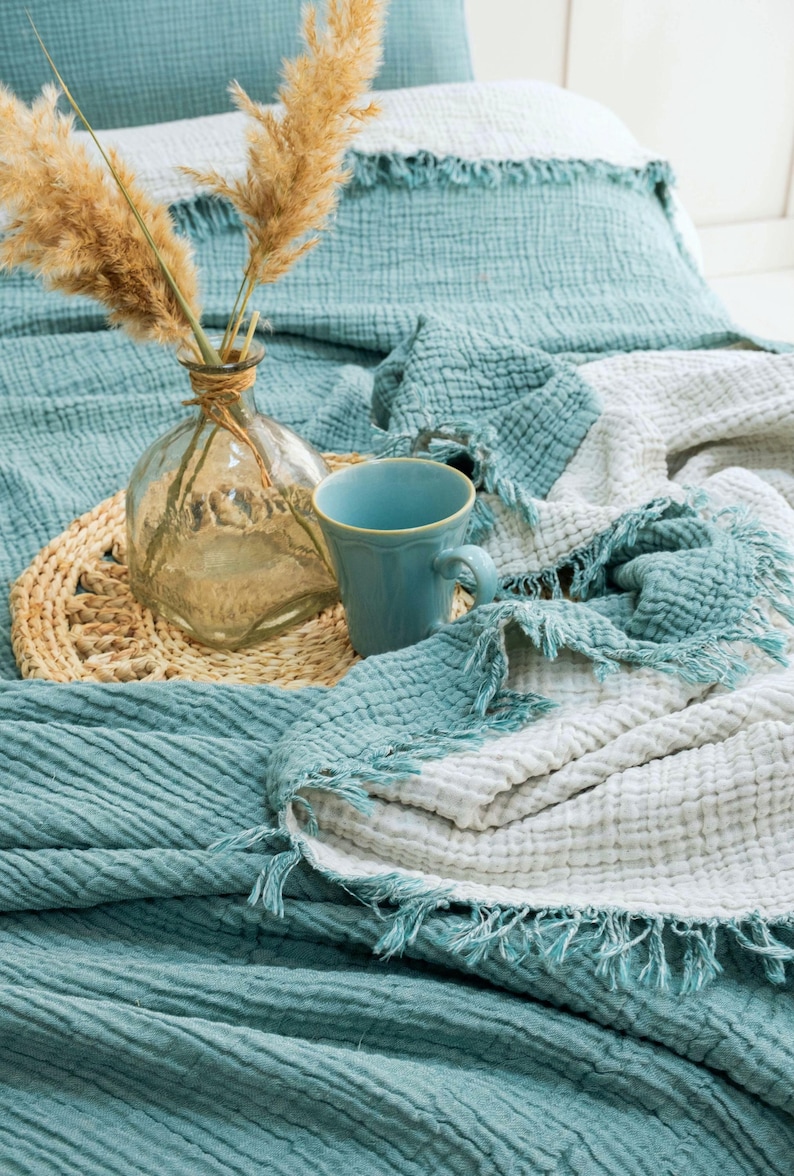 May include: A teal and white crinkled muslin throw blanket with frayed edges, featuring a clear glass vase containing dried pampas grass and a solid teal ceramic mug placed on a round woven straw mat atop the textured fabric surface.
