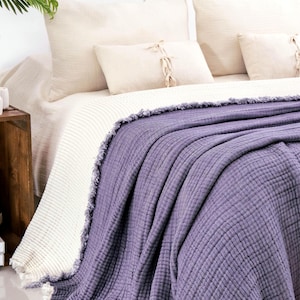 Op de afbeelding: Een dieppaarse, getextureerde mousseline sprei met gerafelde randen gedrapeerd over een bed. De deken heeft een meerlaagse, gekreukelde stofconstructie. Het ligt over een cr&egrave;mekleurige, geribbelde gebreide deken en bijpassende kussens.
