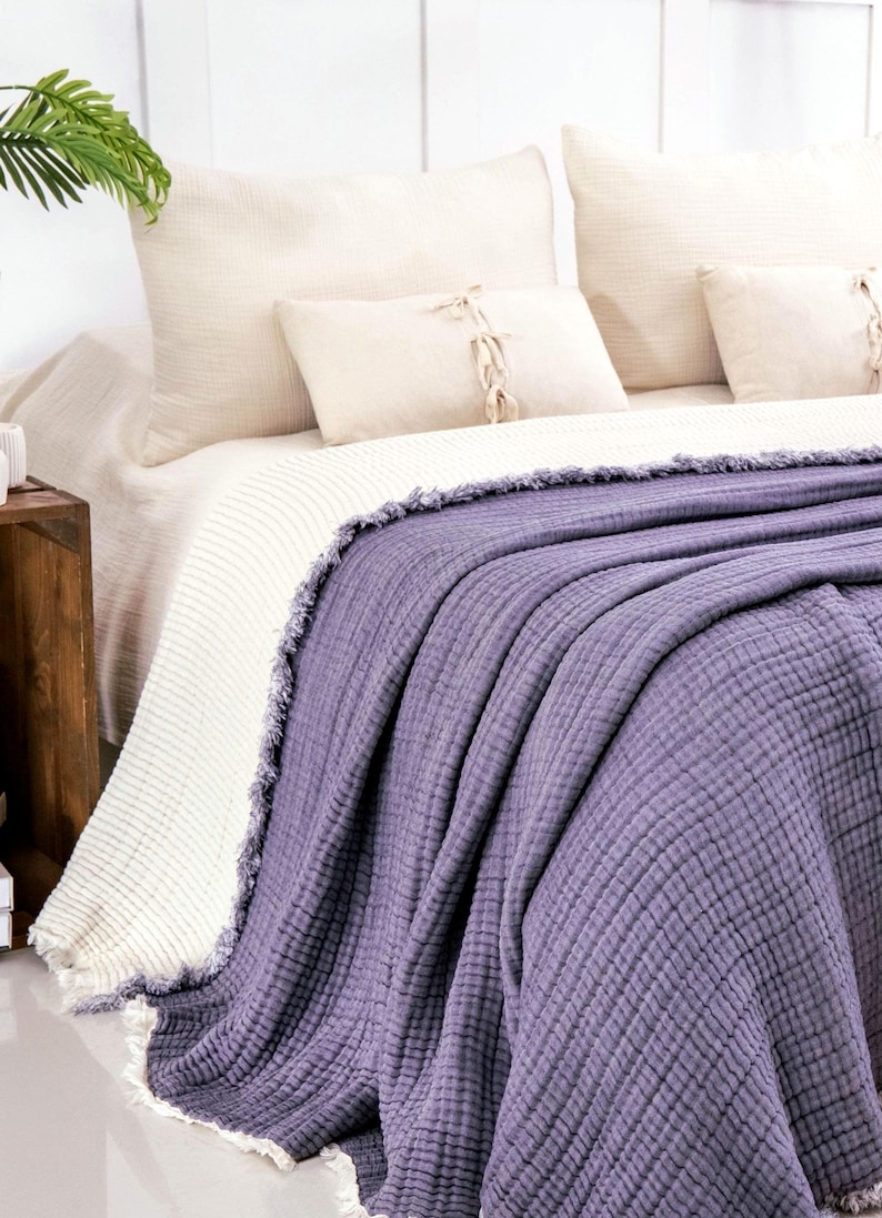 Op de afbeelding: Een dieppaarse, getextureerde mousseline sprei met gerafelde randen gedrapeerd over een bed. De deken heeft een meerlaagse, gekreukelde stofconstructie. Het ligt over een cr&egrave;mekleurige, geribbelde gebreide deken en bijpassende kussens.