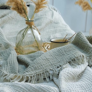 May include: A sage green muslin throw blanket with a textured waffle weave and fringed edges, draped over a surface. A clear glass vase containing dried pampas grass and a small cream-colored enamel mug with a dark rim sit on the blanket.