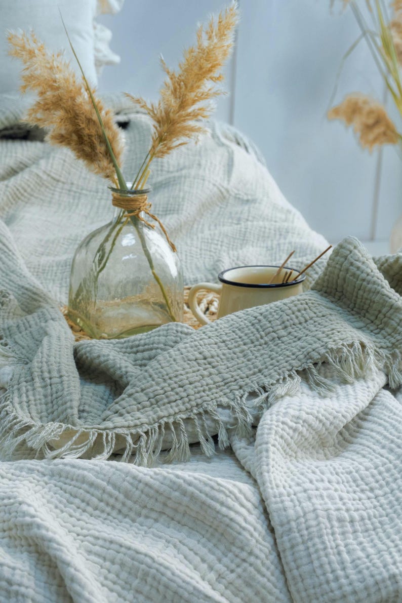 May include: A sage green muslin throw blanket with a textured waffle weave and fringed edges, draped over a surface. A clear glass vase containing dried pampas grass and a small cream-colored enamel mug with a dark rim sit on the blanket.