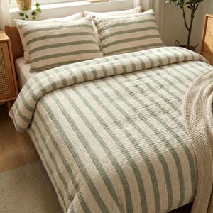 May include: A bed dressed with a cream-coloured duvet cover and matching pillowcases, featuring vertical stripes in a muted green. The bedding is arranged on a wooden bed frame, creating a cosy atmosphere. A neutral-toned blanket is draped over the side.