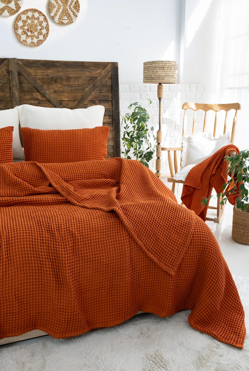 Op de afbeelding: Een bedsprei en bijpassende rechthoekige kussensloop in een gebrande oranje kleur met een wafelstructuur. De stof heeft een duidelijk rasterachtig patroon over het gehele oppervlak. Het beddengoed ligt over een bedframe en toont de diepe, warme kleur en de tastbare, verhoogde textuur van het textiel.