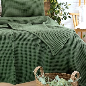 Op de afbeelding: Een donker olijfgroene sprei en bijpassende kussensloop met wafelstructuur. De stof heeft een duidelijk rasterachtig patroon. Op de voorgrond staat een ronde mand van gevlochten zeegras met groene eucalyptusstengels erin.
