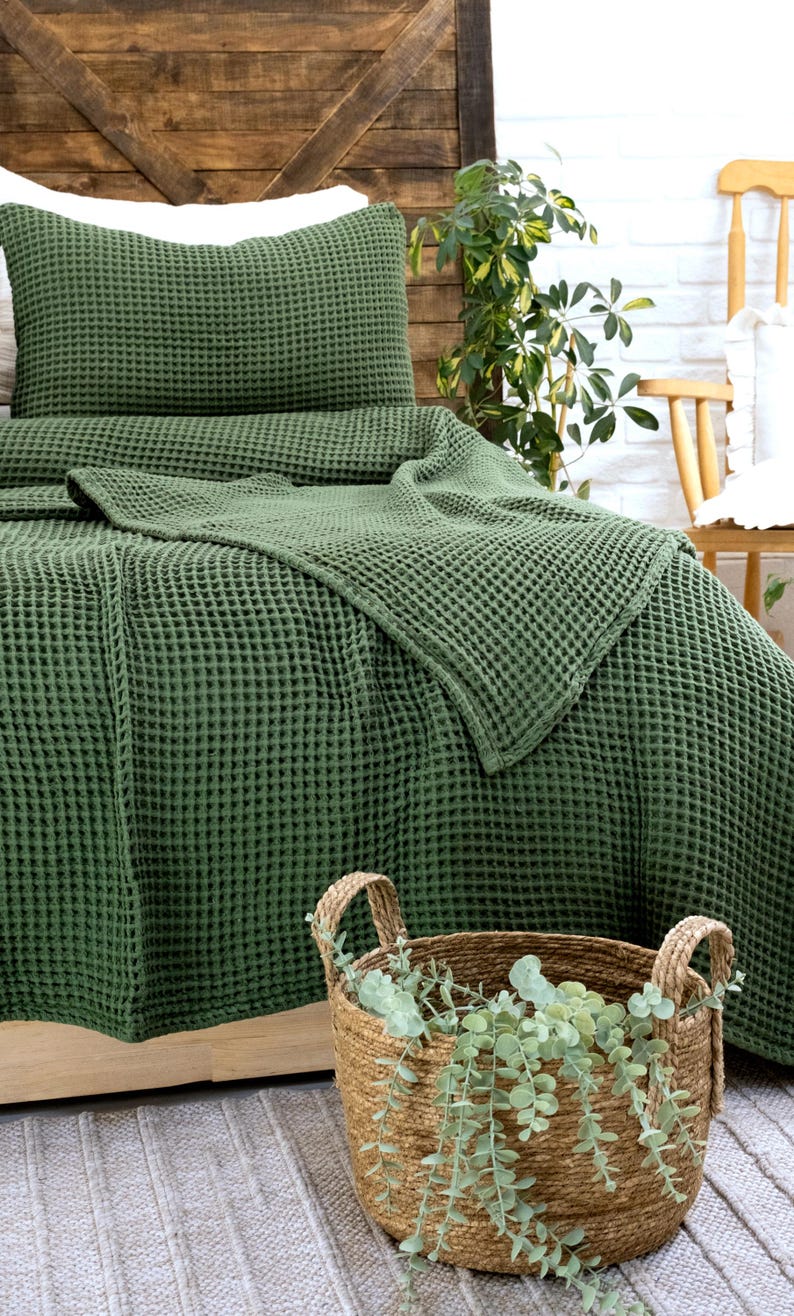 Op de afbeelding: Een donker olijfgroene sprei en bijpassende kussensloop met wafelstructuur. De stof heeft een duidelijk rasterachtig patroon. Op de voorgrond staat een ronde mand van gevlochten zeegras met groene eucalyptusstengels erin.