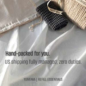 May include: A beige textured fabric bundle tied with a branded ribbon, a small dark gray waffle-weave cloth, and a cream-colored drawstring pouch. The text reads: Hand-packed for you. US shipping fully managed, zero duties. YUMENIA | REFILL ESSENTIALS.