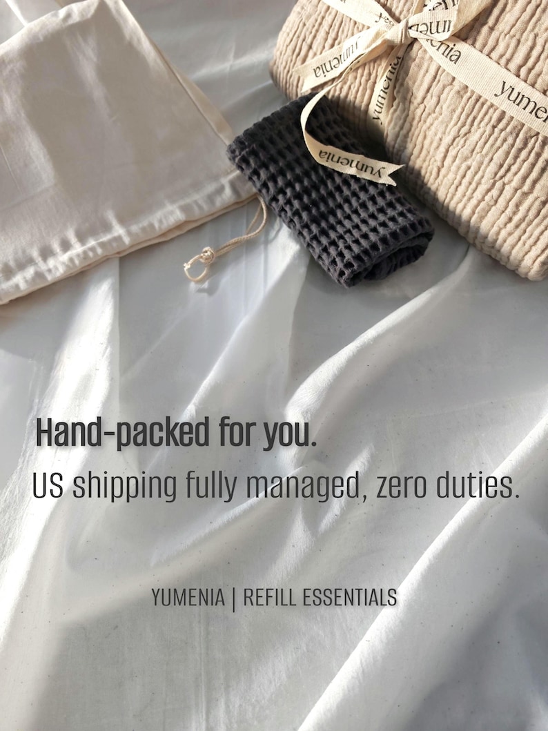 May include: A beige textured fabric bundle tied with a branded ribbon, a small dark gray waffle-weave cloth, and a cream-colored drawstring pouch. The text reads: Hand-packed for you. US shipping fully managed, zero duties. YUMENIA | REFILL ESSENTIALS.