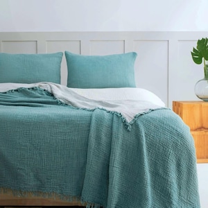 May include: A bed set featuring two teal rectangular pillowcases and a matching teal textured muslin throw blanket with frayed edges, layered over a white sheet. The fabric has a visible grid-like weave pattern throughout the bedding set.