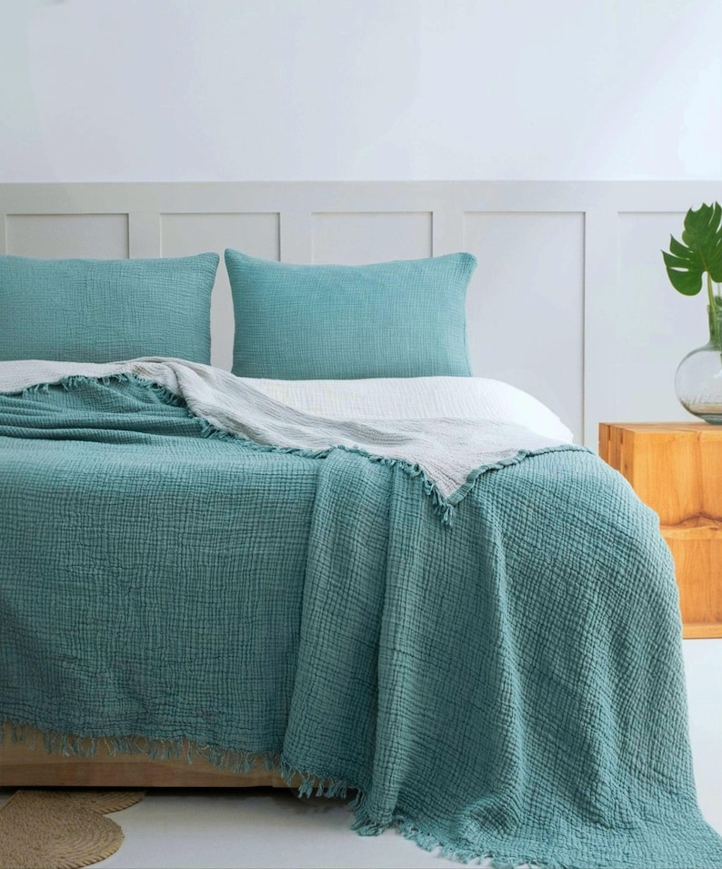 May include: A bed set featuring two teal rectangular pillowcases and a matching teal textured muslin throw blanket with frayed edges, layered over a white sheet. The fabric has a visible grid-like weave pattern throughout the bedding set.
