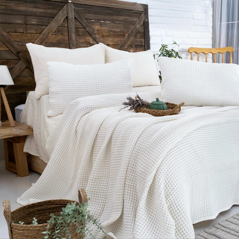 Op de afbeelding: Een witte bedsprei met wafelstructuur en bijpassende kussenslopen. Het beddengoed heeft een textuur met een rasterpatroon. Op het bed staat een klein gevlochten mandje met een groen decoratief object en een bosje gedroogde lavendel.
