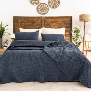Op de afbeelding: Een donkerblauwe bedsprei met wafelstructuur gedrapeerd over een bed, vergezeld van twee bijpassende rechthoekige kussenslopen. Het beddengoed heeft een duidelijke rasterachtige textuur door de hele stof, wat zorgt voor een uniforme uitstraling.