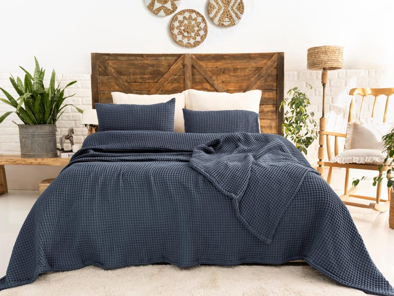 Op de afbeelding: Een donkerblauwe bedsprei met wafelstructuur gedrapeerd over een bed, vergezeld van twee bijpassende rechthoekige kussenslopen. Het beddengoed heeft een duidelijke rasterachtige textuur door de hele stof, wat zorgt voor een uniforme uitstraling.