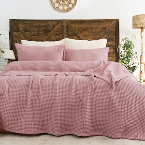 Op de afbeelding: Een oudroze wafelstructuur sprei gedrapeerd over een bed, vergezeld door twee bijpassende rechthoekige kussenslopen. De stof heeft een duidelijke rasterachtige textuur over het gehele oppervlak van de beddengoedset, wat zorgt voor een uniform uiterlijk.