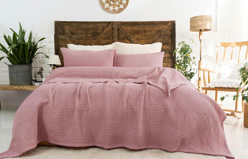 Op de afbeelding: Een oudroze wafelstructuur sprei gedrapeerd over een bed, vergezeld door twee bijpassende rechthoekige kussenslopen. De stof heeft een duidelijke rasterachtige textuur over het gehele oppervlak van de beddengoedset, wat zorgt voor een uniform uiterlijk.