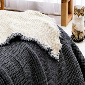 May include: A dark charcoal gray textured waffle-weave blanket layered with a cream-colored waffle-weave blanket. Both blankets feature frayed, fringed edges with a mix of dark and light threads, creating a layered textile appearance.