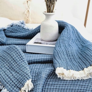 Op de afbeelding: Een blauwe, getextureerde katoenen plaid met een rasterachtig doorgestikt patroon en gerafelde, cr&egrave;mekleurige franjes. De deken is gedrapeerd over een witte rechthoekige doos, die een witte keramische vaas met gedroogde lavendeltakken ondersteunt.