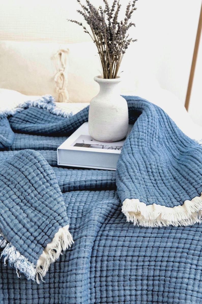 Op de afbeelding: Een blauwe, getextureerde katoenen plaid met een rasterachtig doorgestikt patroon en gerafelde, cr&egrave;mekleurige franjes. De deken is gedrapeerd over een witte rechthoekige doos, die een witte keramische vaas met gedroogde lavendeltakken ondersteunt.