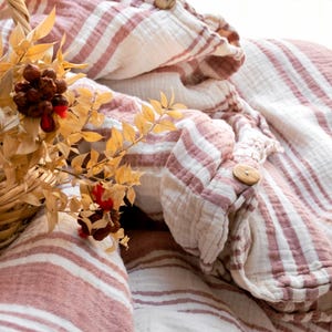 Puede incluir: Una manta de tiro a rayas rosa y blancas con un botón de madera. La manta está hecha de una tela suave y texturizada, perfecta para acurrucarse en el sofá.