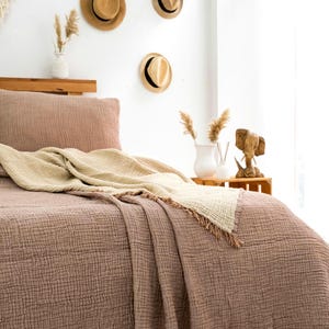 May include: A textured mauve bedspread with a matching pillowcase and a folded cream-colored throw blanket featuring frayed edges. The bedding is made of a crinkled fabric and is draped over a wooden bed frame in a room with neutral decor.