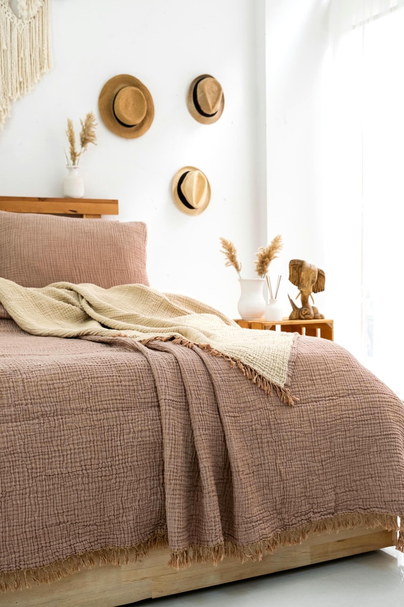 May include: A textured mauve bedspread with a matching pillowcase and a folded cream-colored throw blanket featuring frayed edges. The bedding is made of a crinkled fabric and is draped over a wooden bed frame in a room with neutral decor.