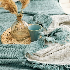 May include: A teal and white reversible muslin throw blanket with frayed edges, featuring a crinkled texture. A clear glass vase containing dried pampas grass and a light blue ceramic mug sit on a round woven placemat atop the folded blanket.