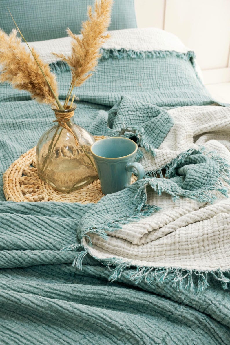 May include: A teal and white reversible muslin throw blanket with frayed edges, featuring a crinkled texture. A clear glass vase containing dried pampas grass and a light blue ceramic mug sit on a round woven placemat atop the folded blanket.