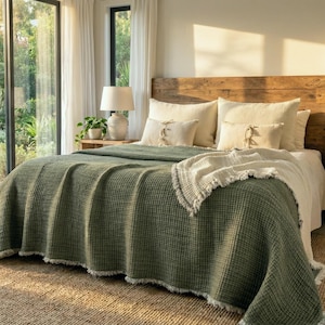 May include: A bedroom featuring a bed with a textured olive green waffle-weave blanket, cream-coloured pillows, and a wooden headboard. Two bedside tables with lamps are positioned on either side of the bed, which is set on a woven jute rug.