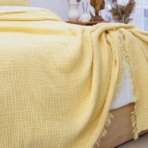 May include: A light yellow muslin blanket with a textured, crinkled surface and frayed edges. The fabric is draped over a bed, showing a soft, layered appearance with visible stitching details along the border of the textile.