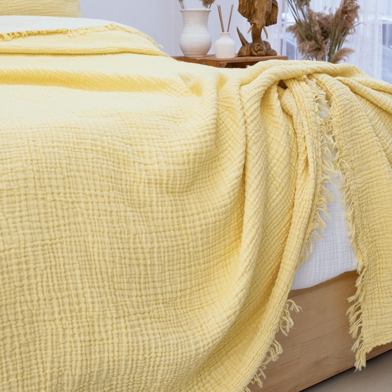 May include: A light yellow muslin blanket with a textured, crinkled surface and frayed edges. The fabric is draped over a bed, showing a soft, layered appearance with visible stitching details along the border of the textile.