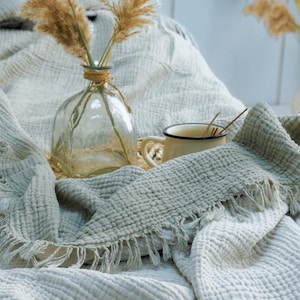 May include: A sage green muslin throw blanket with a textured, crinkled surface and fringed edges. The blanket is draped near a clear glass vase containing dried pampas grass and a small cream-colored mug with a dark rim and a metal stirring spoon.