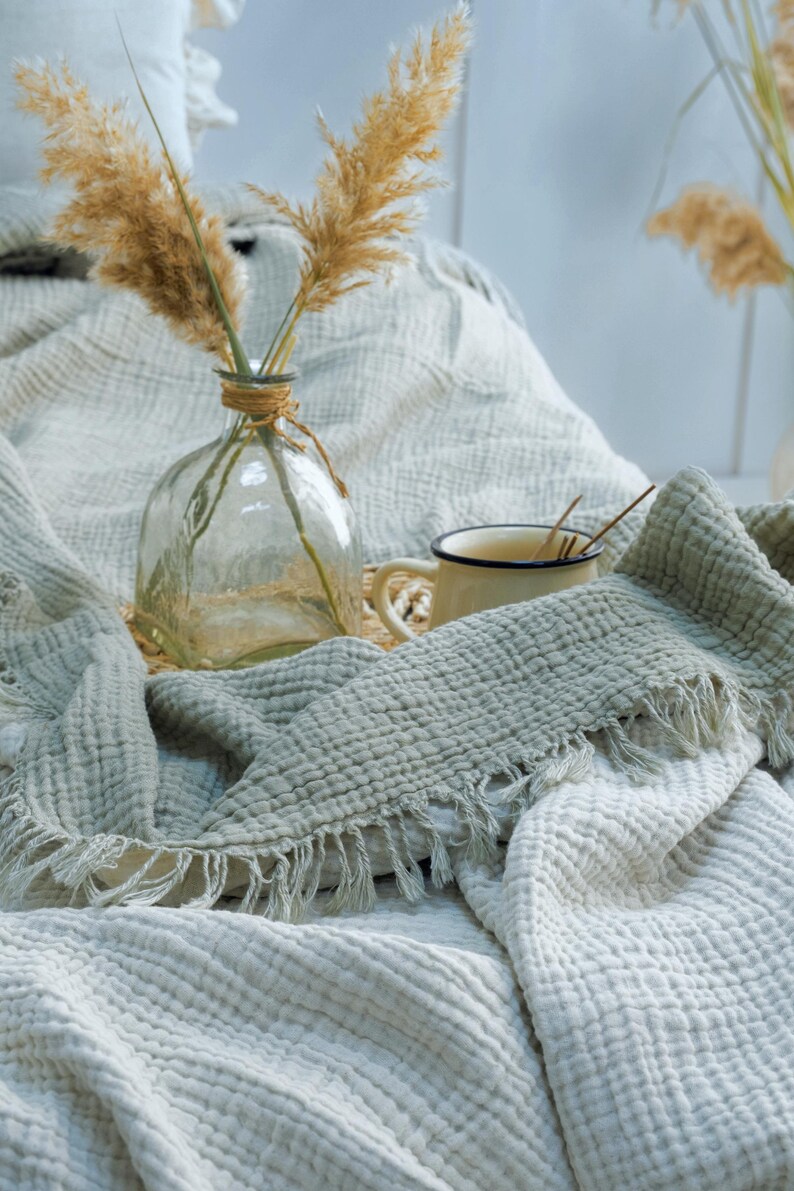 May include: A sage green muslin throw blanket with a textured, crinkled surface and fringed edges. The blanket is draped near a clear glass vase containing dried pampas grass and a small cream-colored mug with a dark rim and a metal stirring spoon.