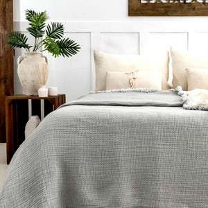 May include: A gray throw blanket with a fringed edge, folded on a bed with white pillows. The blanket has a waffle-like texture.