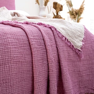 May include: A textured, crinkled cotton bedspread in a deep mauve color, featuring a matching pillowcase and a folded-back cream-colored layer. The edges of the fabric are finished with a fringe detail, creating a soft, layered appearance on the bed.
