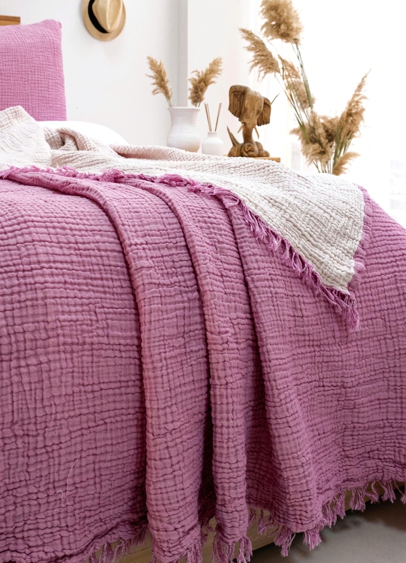 May include: A textured, crinkled cotton bedspread in a deep mauve color, featuring a matching pillowcase and a folded-back cream-colored layer. The edges of the fabric are finished with a fringe detail, creating a soft, layered appearance on the bed.