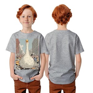 Puede incluir: Una camiseta gris con un gran gráfico de un ganso en un entorno urbano. El ganso es blanco con un cuello largo y es el punto focal. El fondo muestra edificios y personas. La camiseta la lleva un niño.
