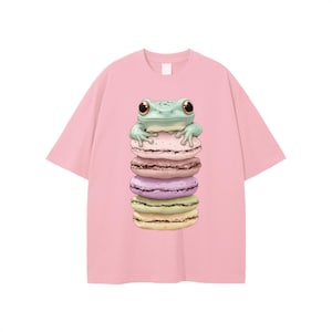May include: A light pink t-shirt featuring a cartoon frog perched atop a stack of colorful macarons. The frog is light green with large brown eyes. The macarons are pink, purple, and yellow.