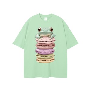 Peut inclure: T-shirt vert clair avec un motif de grenouille de dessin animé. La grenouille est assise sur une pile de macarons roses, violets et beiges. La grenouille a de grands yeux. Le t-shirt est de coupe ample.