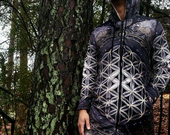 Puede incluir: Una chaqueta con capucha y cremallera de manga larga con una base negra y un patrón geométrico repetitivo de flor de la vida en blanco y dorado. La prenda incluye una capucha con forro interior claro, una cremallera frontal completa y dos bolsillos.